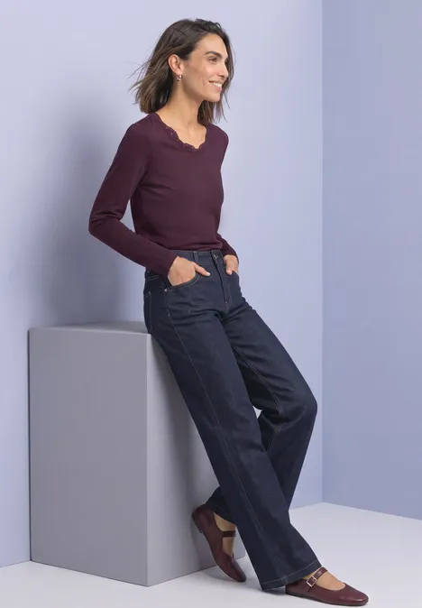 Langarm Shirt mit Rundhals und Spitze rustic rouge Frau lehnt an grauem Podest, trägt weinrotes Langarmshirt, weite dunkle Jeans und braune Schuhe vor hellem Hintergrund.