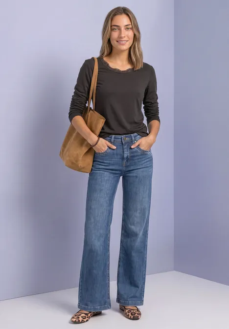 Frau steht vor neutralem Hintergrund, trägt schwarzen Pullover, weite Jeans, Sandalen und eine beige Schultertasche.