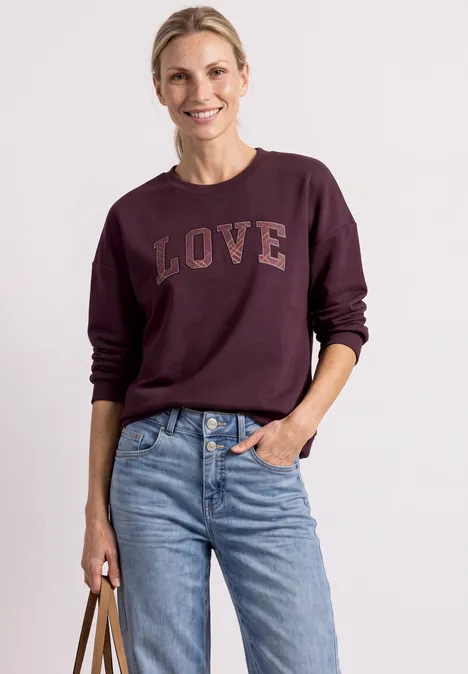 Frau in weinrotem Sweatshirt mit LOVE-Print, Jeans und Handtasche, lächelt.