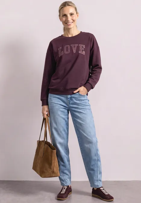 Lächelnde Frau vor hellem Hintergrund in weinrotem LOVE-Sweatshirt, Jeans, braunen Sneakern und Tasche.