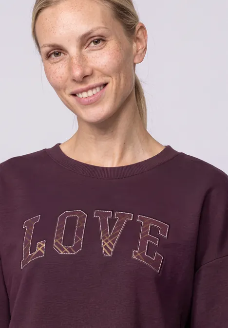 Eine lächelnde Frau mit hellblondem Haar im Pferdeschwanz, trägt ein violettes Sweatshirt mit dem Schriftzug 'LOVE'.