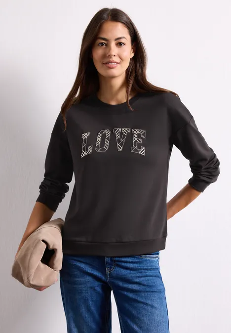Langarm Shirt mit Wording Artwork black coffee Frau trägt schwarzen Pullover mit LOVE-Schriftzug, kombiniert mit blauen Jeans und hält eine beige Jacke.