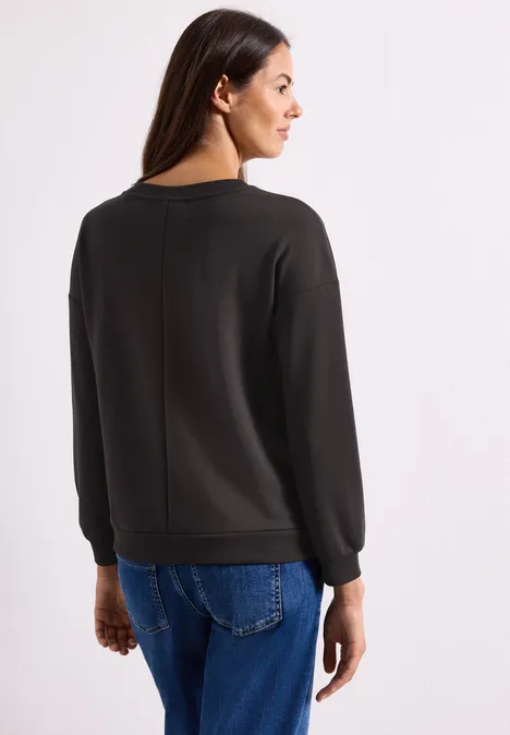 Langarm Shirt mit Wording Artwork black coffee Frau steht mit dem Rücken zur Kamera, trägt schwarzen Pullover und blaue Jeans vor weißem Hintergrund.