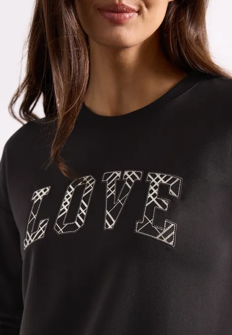 Langarm Shirt mit Wording Artwork black coffee Eine Frau trägt ein schwarzes Sweatshirt mit dem Schriftzug LOVE in weißem Grafik-Stil.