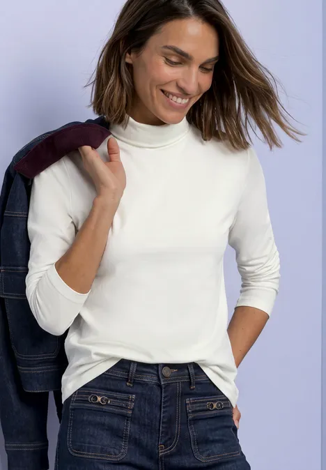 Lächelnde Frau im weißen Rollkragenpullover und Jeans hält Jacke über der Schulter vor lila Hintergrund.
