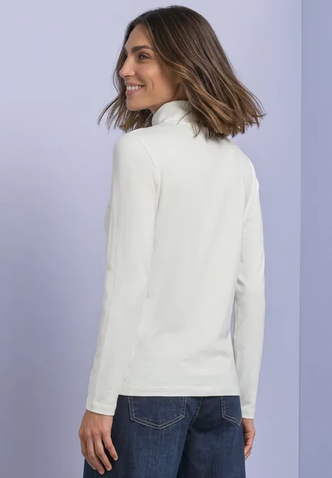 Eine lächelnde Frau in weißem Rollkragenpullover blickt über die Schulter, vor lila Hintergrund.