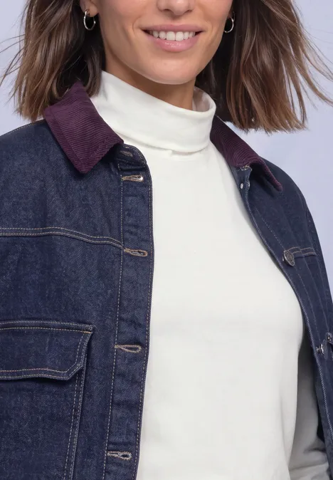 Eine Frau trägt eine dunkle Jeansjacke mit burgunderfarbenem Kragen und einen weißen Rollkragenpullover.