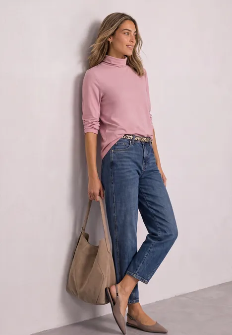 Eine Frau lehnt an einer Wand, trägt rosa Pullover, blaue Jeans, Wildleder-Flats und eine große Tasche.