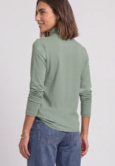 Frau in olivgrüner Rollkragenlongsleeve und Jeans, schaut lächelnd seitlich.