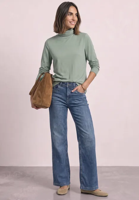 Frau mit grünem Rollkragenpullover, geraden blauen Jeans und brauner Tasche lächelt vor hellem Hintergrund.