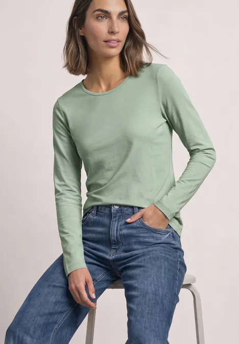 Lot de 2 t-shirts à manches longues et col rond shadow teal/eucalyptus green Eine Frau sitzt lässig auf einem Hocker, trägt ein hellgrünes Langarmshirt und blaue Jeans, eine Hand in der Tasche.