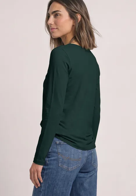 Lot de 2 t-shirts à manches longues et col rond shadow teal/eucalyptus green Frau trägt dunkelgrünes Langarmshirt und Jeans, blickt über die Schulter vor hellem Hintergrund.
