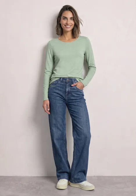 Lot de 2 t-shirts à manches longues et col rond shadow teal/eucalyptus green Eine Frau trägt ein grünes Langarmshirt, weite Jeans und weiße Schuhe vor hellem Hintergrund.