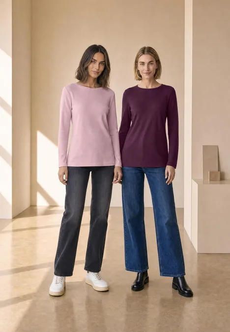 Twee vrouwen in effen truien en jeans poseren in een lichte, minimalistische ruimte.