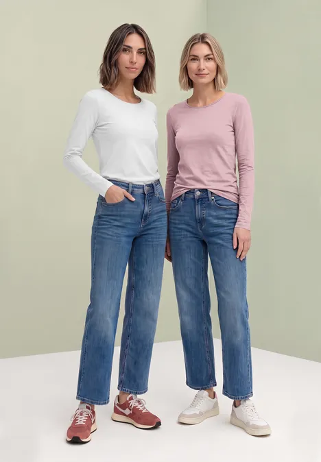 Zwei Frauen in Jeans und langärmligen Shirts posieren vor hellgrünem Hintergrund.
