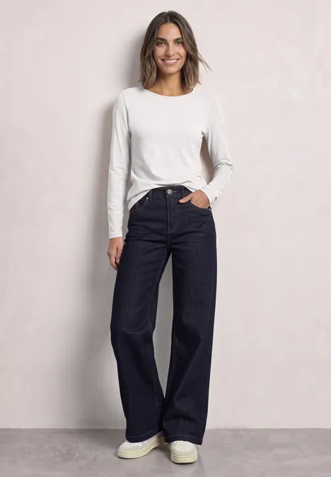 Eine Frau trägt ein weißes Langarmshirt und weite, dunkle Jeans und lächelt vor hellem Hintergrund.