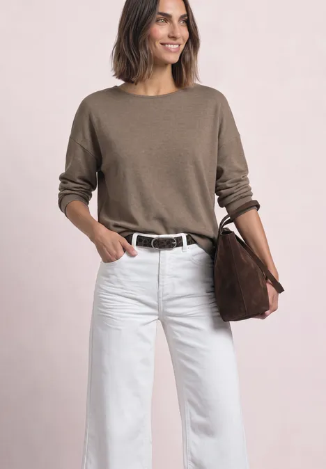 Eine lächelnde Frau trägt einen beigen Pullover, weiße Hose und hält eine braune Tasche.