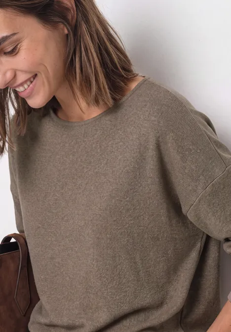 Lächelnde Frau in lockerem khakifarbenem Pullover hält braune Tasche, lässiger Casual-Look.