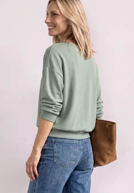 Eine blonde Frau im graugrünem Pullover und Jeans, lächelnd mit einer braunen Clutch in der Hand, seitlich stehend.