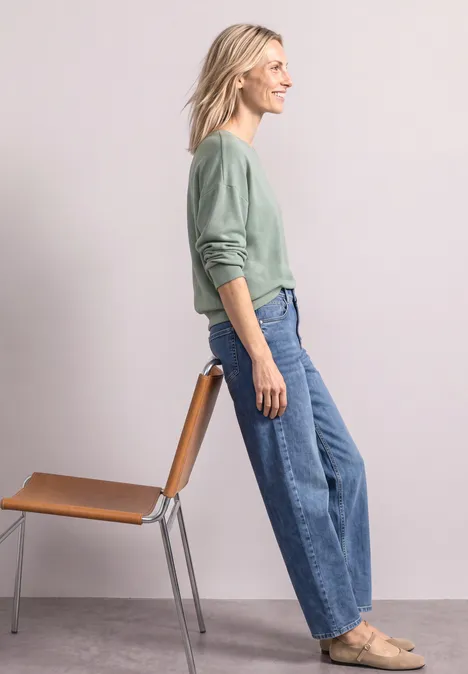 Dame im grünen Pullover lehnt an Stuhl, trägt weite Jeans und flache Schuhe
