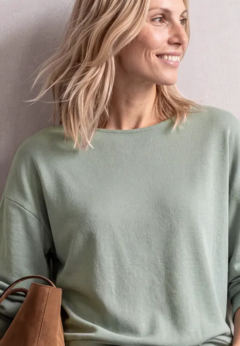 Lächelnde Frau mit blondem Haar in grünem Pullover, seitlich blickend, mit Schultertasche.