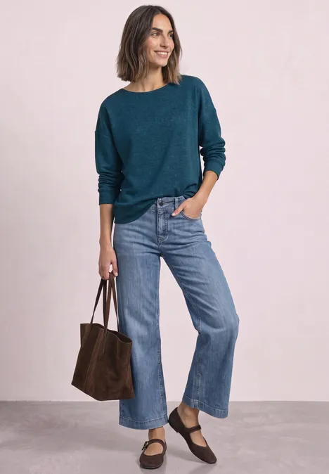 Frau in türkisfarbenem Pullover und weiten Jeans hält eine braune Tasche. Sie lächelt leicht.
