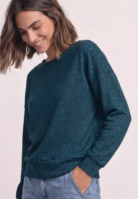 Frau lächelt in einem dunkelgrünen, leicht glitzernden Pullover mit langen Ärmeln und Taschen.