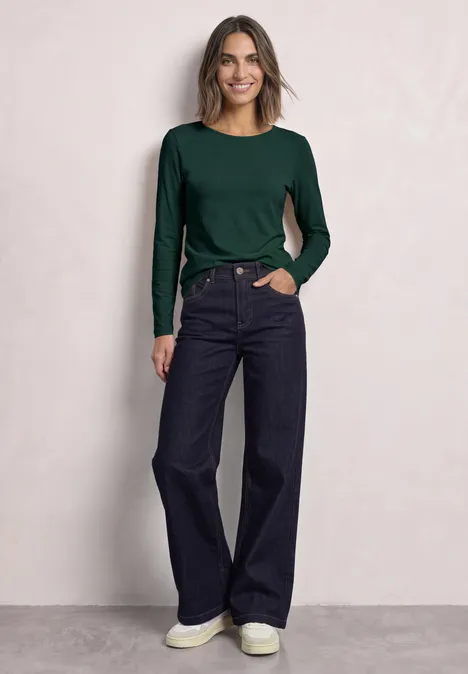 Lot de 2 t-shirts à manches longues et col bateau shadow teal/eucalyptus green Frau mit grünem Langarmshirt, weitem Dunkelblau-Jeans und weißen Sneakern vor neutraler Wand.