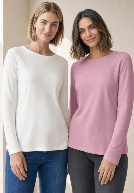 Zwei lächelnde Frauen in langärmligen Pullovern (weiß und rosa) und Jeans vor neutralem Hintergrund.