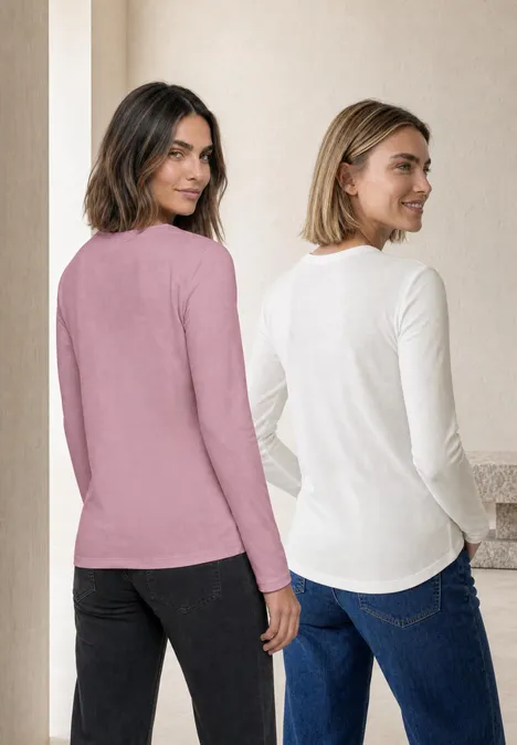 Zwei Frauen stehen Rücken an Rücken und tragen langärmelige Oberteile in Rosa und Weiß mit Jeans.