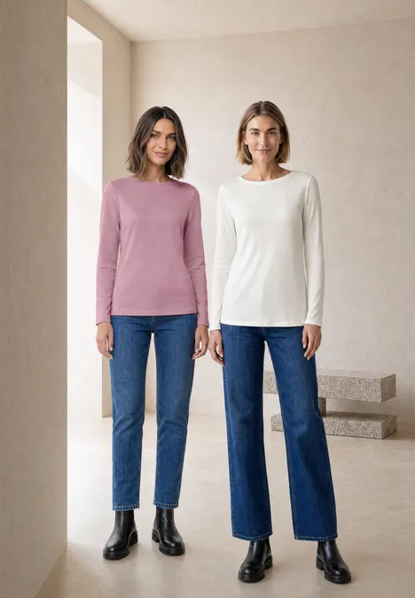 Zwei Frauen stehen vor hellem, minimalistischem Hintergrund und tragen Jeans und Langarmshirts.