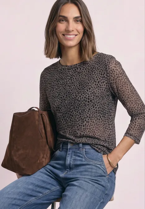Frau mit braunem Bob, in durchsichtiger, gepunkteter Bluse, blauen Jeans und brauner Tasche, lächelnd.