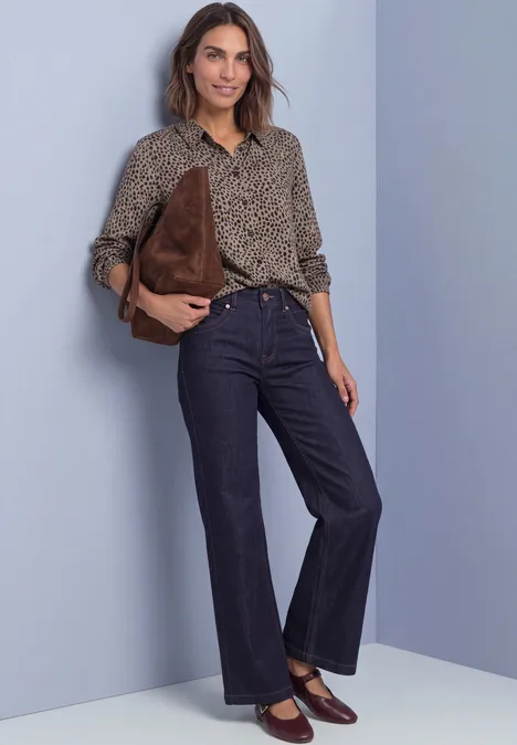 Eine Frau in geblümter Bluse und dunklen Jeans hält eine braune Tasche vor lila Hintergrund.