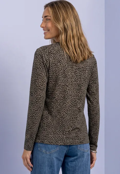 Frau in gemustertem Langarmshirt und Jeans, von hinten leicht seitlich blickend, vor hellem Hintergrund.