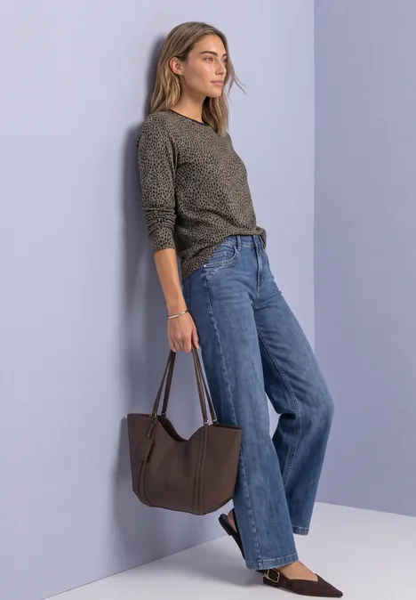 Frau lehnt an lila Wand, trägt graues Oberteil, weite Jeans und hält eine braune Tasche.