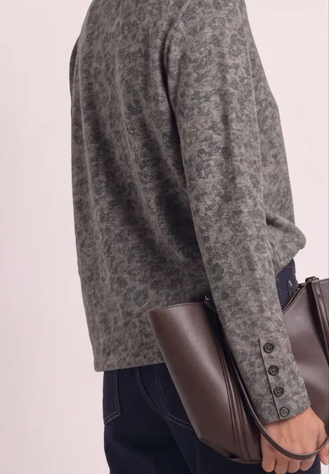 Person in grauem gemustertem Blazer, dunkler Jeans, hält eine braune Ledertasche.