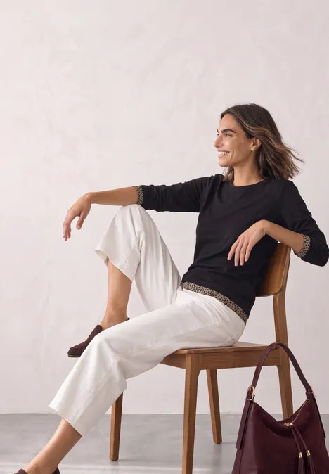 Vrouw in een zwarte trui en witte broek, glimlachend zittend op een houten stoel met tas ernaast.