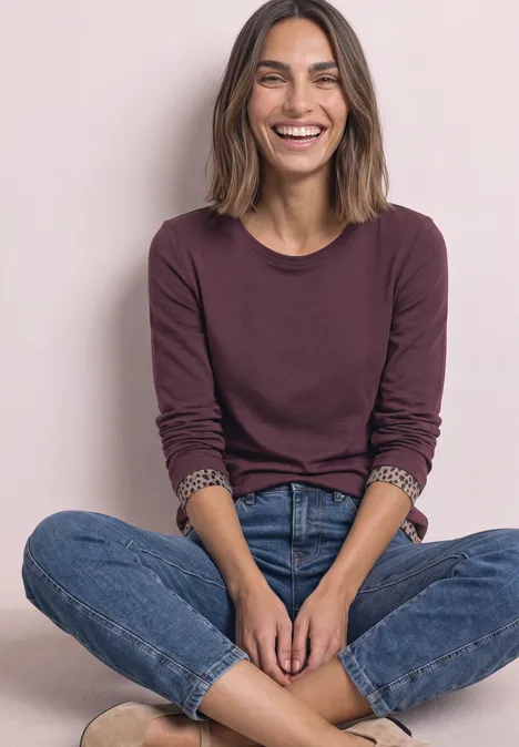 Eine lächelnde Frau in Jeans und weinrotem Langarmshirt sitzt im Schneidersitz vor hellem Hintergrund.