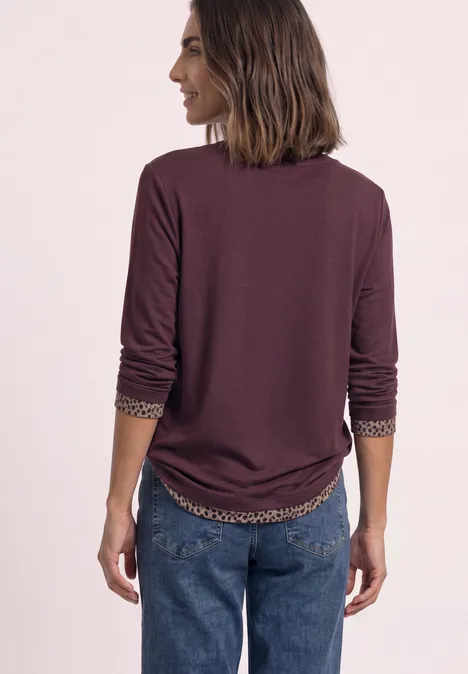 Rückenansicht einer Frau in bordeauxroter, langärmliger Bluse mit gemusterter Innenmanschette und blauen Jeans.