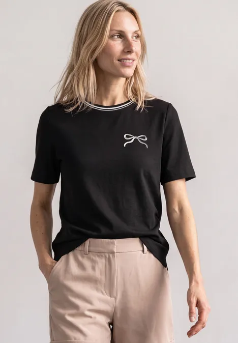 Frau in schwarzem T-Shirt mit weißem Schleifen-Print, beiger Hose, Hände in den Taschen, blickt zur Seite.