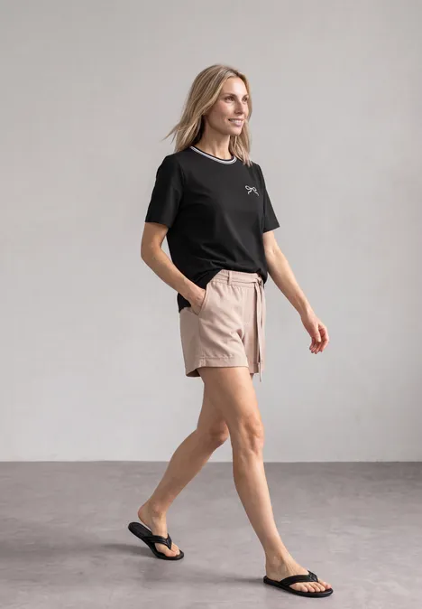 Frau in schwarzem Shirt, beigen Shorts und Flipflops schreitet gelassen.