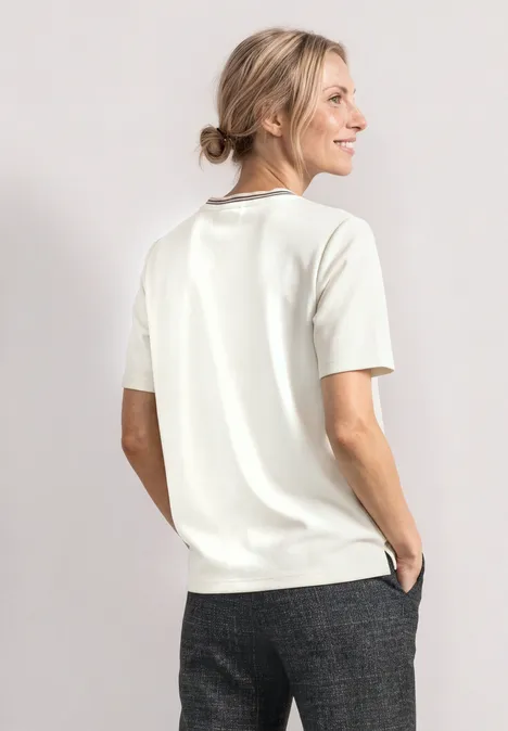 T-shirt à col rond avec détail brodé off white Frau in weißem Kurzarm-Pullover und grauer Hose, seitlich stehend und lächelnd vor hellem Hintergrund.