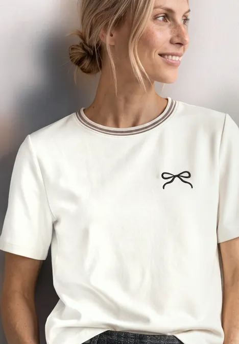 T-shirt à col rond avec détail brodé off white Eine lächelnde Frau mit blondem Dutt trägt ein weißes T-Shirt mit dünnem Ringel-Kragen und kleinem Schleifen-Print.