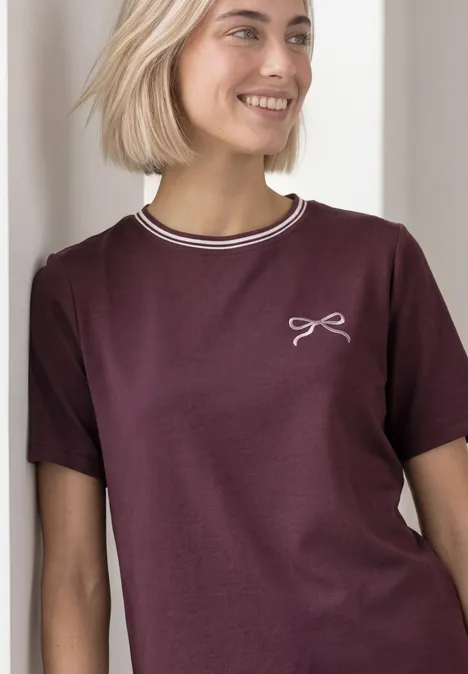 Eine junge Frau in einem burgunderroten T-Shirt mit weißem Kragenrand und kleinem Schleifen-Motiv.