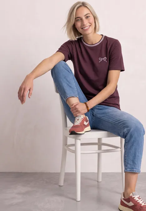 Eine junge Frau sitzt auf einem weißen Stuhl, lächelnd in Jeans, bordeauxrotem T-Shirt und Sneakern.