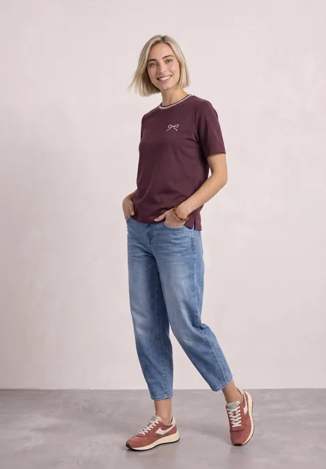 Frau mit blondem Haar in weinrotem T-Shirt, blauen Jeans und Sneakers, lächelnd vor hellem Hintergrund.
