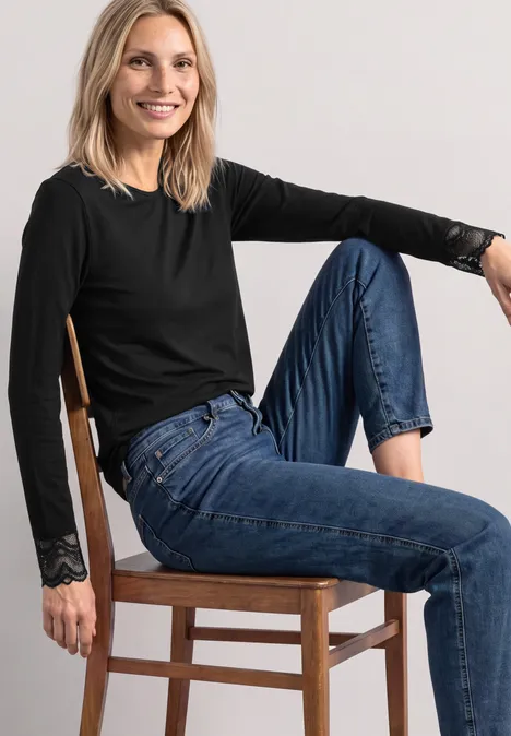 Langarm Shirt mit Rundhals und Spitze Black Eine lächelnde Frau in schwarzem Langarmshirt und blauen Jeans sitzt auf einem Holzstuhl vor hellem Hintergrund.