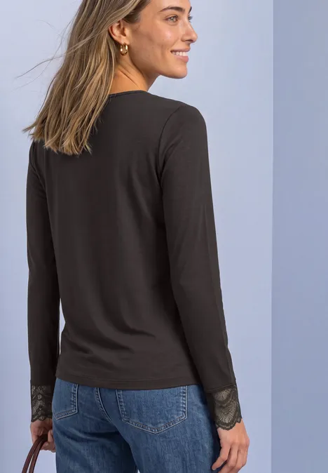 Eine lächelnde Frau von hinten in schwarzem Langarmshirt mit Spitzenärmeln und blauer Jeans.