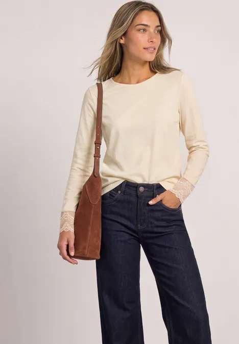 Frau mit cremefarbenem Langarmshirt mit Spitze, dunkelblauer Jeans und brauner Umhängetasche.