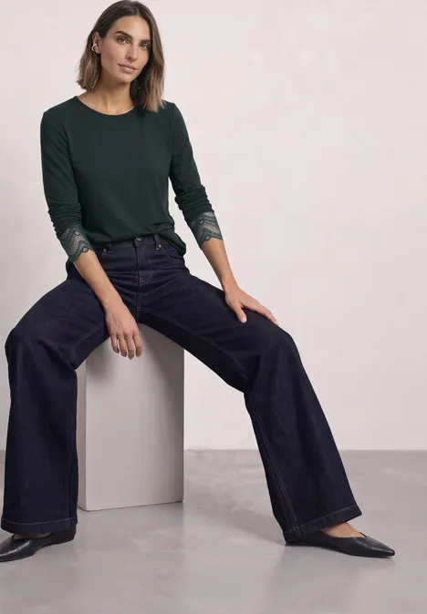 Langarm Shirt mit Rundhals und Spitze stormy green Eine Frau in dunklem Langarmshirt und weitem Jeans sitzt lässig auf einem weißen Würfel.